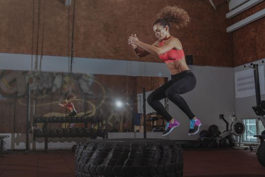 Beautiful fit sportswoman exercising at crossfit box gym, jumping high on a huge tire. Energectic female crossfit athlete working on her endurance and agility, copy space