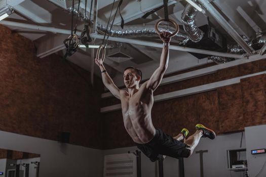 Low angle shot of a male crossfit athlete exercising on gymnastic rings. Shirtless strong sportsman working out on gymnastic rings at crossfit box gym