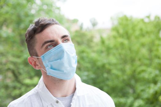 Young man wearing medical protective mask looking away dreamily, enjoying warm day outdoors. Coronavirus pandemic concept