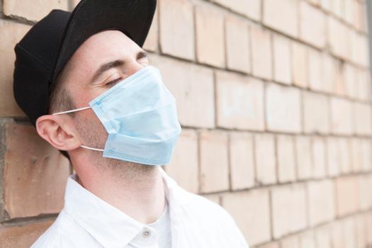 Stylish man leaning on a brick wall in the city, wearing protective medical mask during pandemic