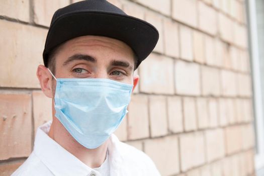 Close up of an attractive stylish man wandering in the city, relaxing near brick wall wearing medical mask on his face