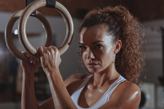 Close up of a beautiful crossfit female athlete looking to the camera concentrated, holding gymnastic rings. Attractive athletic woman preparing for her gymanstic rings workout