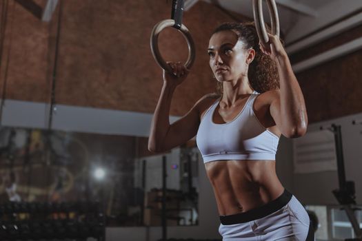 Low angle shot of a beautiful curly haired sportswoman with perfect fit body preparing to exercise on gymnastic rings. Attractive female crossfit athlete resting after workout, copy space