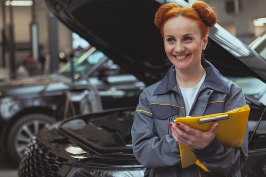 Beautiful red haired woman working at car service station, looking away joyfully, filling papers on clipboard. Attractive female mechanic enjoying working at her garage. Female equality, professions concept