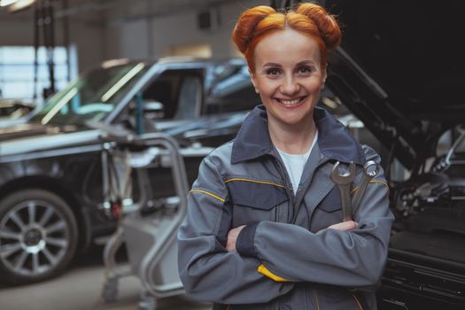 Beautiful happy female car technician in grey uniform posing proudly at her car service station, copy space. Charming woman enjoying working at the garage, repairing modern cars. Professionalism, experience concept