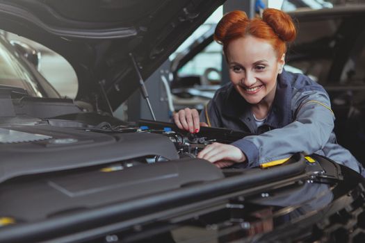 Attractive cheerful red haired female mechanic smiling joyfully, repairing a car at the garage. Lovely woman automobile technician repairing car at the workshop, copy space