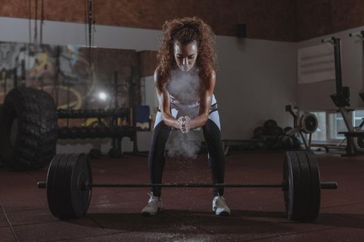 Attractive curly haired female crossfit athlete chalking her hands before lifting heavy barbell. Fit and toned sportswoman using magnesium at crossfit box gym, preparing for weighlifting