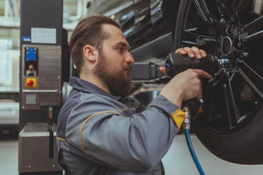 Handsome bearded car mechanic servicing an automobile at his workshop, torquing the lug nuts. Attractive male technician in grey uniform repairing wheels of a SUV vehicle. Safety, insurance concept