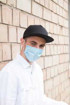 Vertical shot of a cheerful man laughing, wearing medical protective mask, leaning on a brick wall