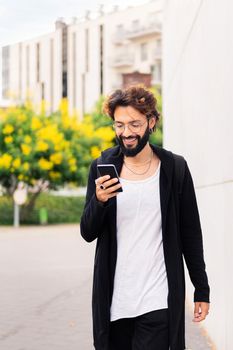smiling young man with beard walking happy by the city using a mobile phone, concept of urban lifestyle and technology of communication, copy space for text