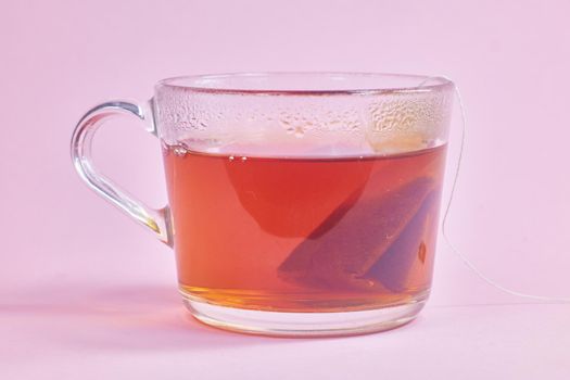 the tea bag is brewed in a transparent glass cup on a pink background.
