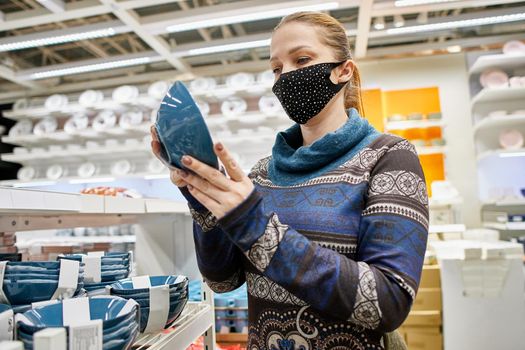 a woman in a protective mask chooses dishes and kitchen accessories in a supermarket, life with protection from infection in the new realities.