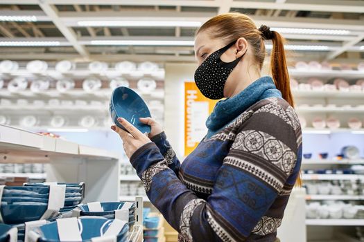 a woman in a protective mask chooses dishes and kitchen accessories in a supermarket, life with protection from infection in the new realities.