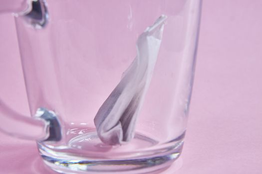 tea bag in an empty transparent glass cup on a pink background.