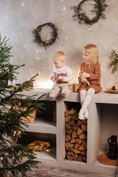 Little brother and sister play on Christmas eve in a beautiful house decorated for the New Year holidays. Children are playing with a Christmas gift. Scandinavian-style interior with live fir trees and a wooden staircase.