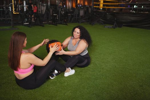 Top view shot of two plus size sportswomen exercising with medicine ball on gym floor, copy space. Overweight female friends doing abs workout, using medicine ball