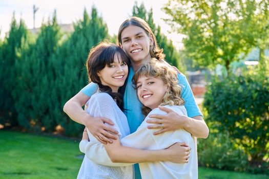 Portrait of three happy teenage friends hugging looking at camera. Guy and two girls posing outdoor together. Friendship, youth, lifestyle, young people concept