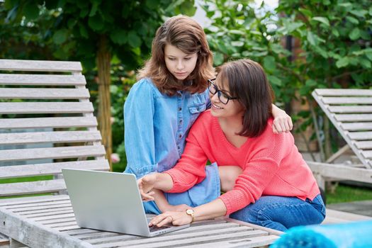 Middle aged mom and preteen daughter looking at laptop together, outdoor on armchairs in backyard. Parent and child hugging using laptop to study leisure shopping