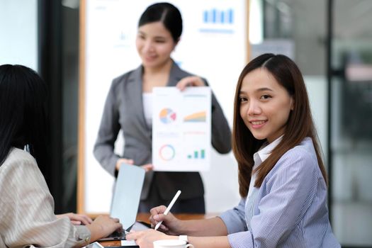 Business. Team meetings. A group sitting down around a table. One person in the foreground. A confident businesswoman.