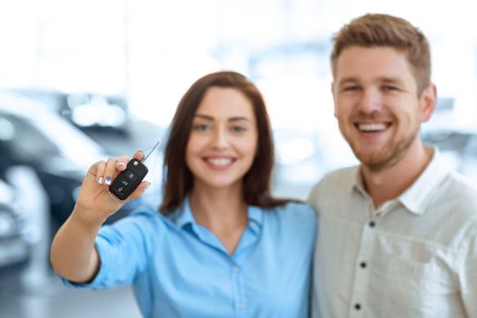 A key to the new chapter. Selective focus on a car key beautiful happy woman is holding hugging with her handsome husband in a car salon