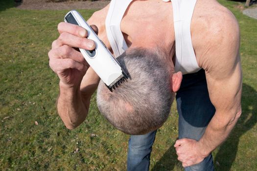 a gray-haired middle-aged man shaves his hair with a clipper in a garden on a green lawn, High quality photo