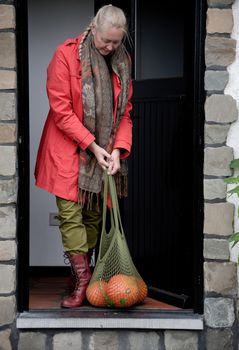 A middle-aged woman in a red cloak with a net of orange pumpkins opens the door to the house, and a kitten meets her on the threshold. High quality photo