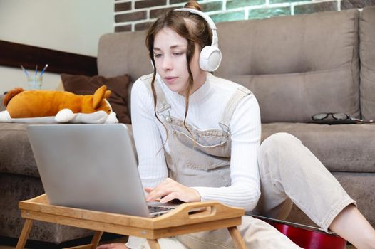 Smiling young woman with laptop watching content or learning while sitting at home in the living room on the floor in casual clothes and headphones.