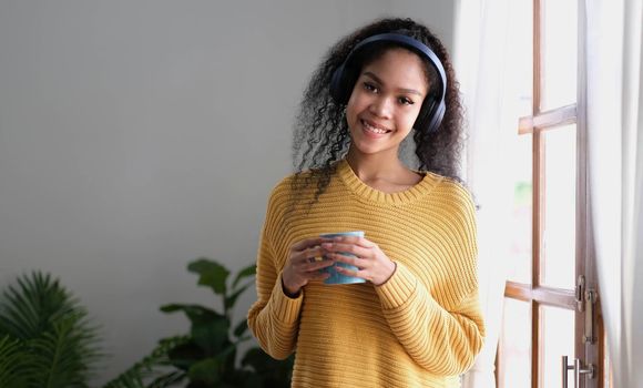 asian woman in headphones holding cup of coffee while standing near window at home.