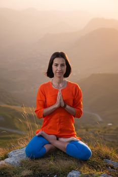 Yogini in the lotus position in full face sits on a cliff against the backdrop of the sunset sky with mountains, prays.