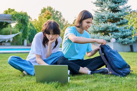 Two teenage friends of students sitting on grass with laptop, in backyard, guy and girl 18 years old study together. Friendship, youth, technology, high school, college, lifestyle concept