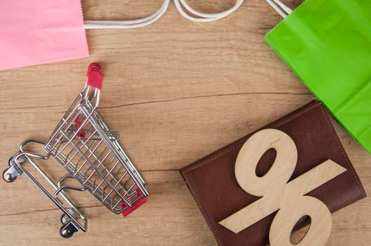 E-commerce online shopping idea concept. Trolley cart with colorful paper shopping bags and leather wallet with sign percentage on wooden table. Customer buying product/service via the internet. Top view. Copy space.