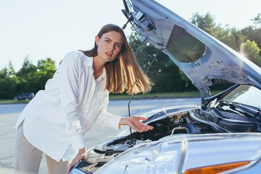 An annoyed woman looks at the camera, depressed near a broken car