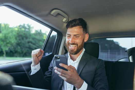 Happy and successful male businessman car passenger reads the news from the phone, rejoices in the victory and the news received