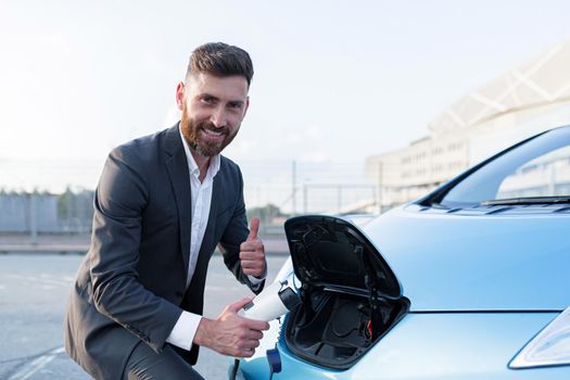 Male businessman charges and refills his electric car, looks happy at the camera and smiles, shows approving thumbs up