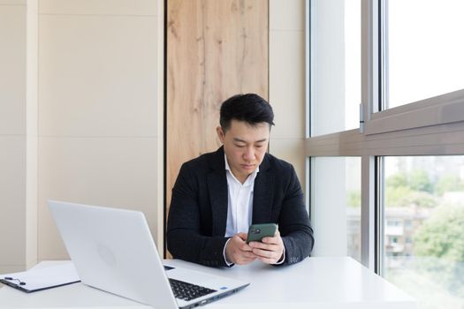 Serious asian business businessman uses cellphone to communicate with colleagues while sitting in modern office