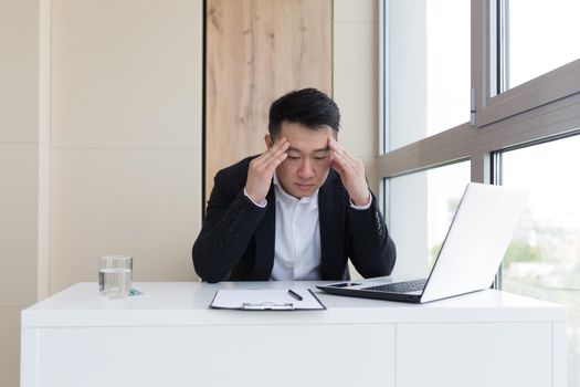 young asian office worker suffering from headache at work in the office drinks medicine pill with water. Sick man in a suit at the computer indoors with severe pain ache uses the drug in workplace