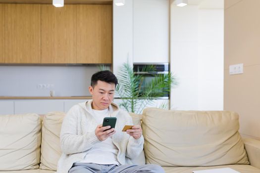 young happy asian man shopping online in internet store using mobile phone and credit card. male enters data of paying for services or tickets in the application sitting on couch at home. easy app