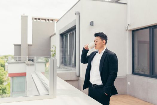 Young successful Asian businessman in a business suit drinking tea or coffee standing on the balcony or terrace of a modern office center