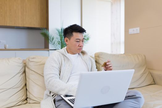 young happy asian man shopping online in internet store using laptop computer and credit card. male enters data of paying for services or tickets in application sitting on couch at home living room