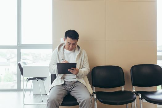 young asian male in waiting room for interview or meeting holding paper while sitting at chair fills out a resume questionnaire in office