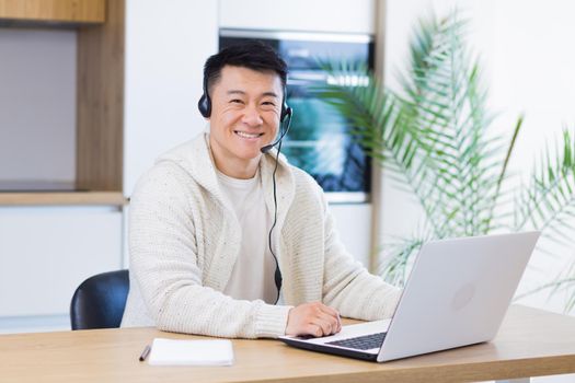 Young asian man with headset looks at camera and smiles sitting at home in the kitchen and working on laptop