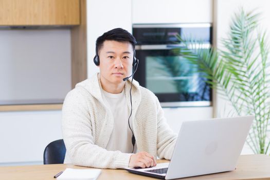Young asian man with headset looks at camera and smiles sitting at home in the kitchen and working on laptop