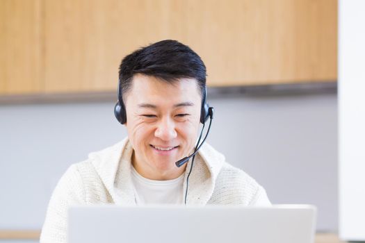 young asian man freelancer sitting at home at office desk with headset and talking online on video link via laptop in room or kitchen. The male indoors in casual clothes does the work and talks