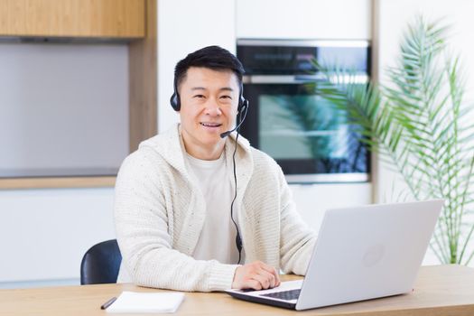 Young asian man with headset looks at camera and smiles sitting at home in the kitchen and working on laptop