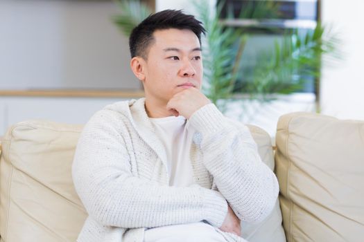 headshot of a young asian man holding his head with a severe headache at home in a room on the couch. Up close, the casual male massages the temples and suffers from chronic pain and fatigue. Closeup