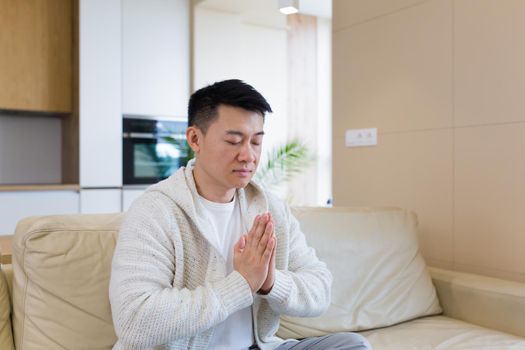 young asian man praying at home alone. Male folding pencil arms and meditating in apartment room. Christian religion traditional spiritual for their hope and believed god forgievness at house