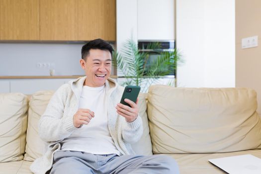 Happy young asian man at home looking at cellphone, with emotion of winner or victory, financial promotion sports betting. The man happily exclaims while playing the game. Excited with joy, celebrating success