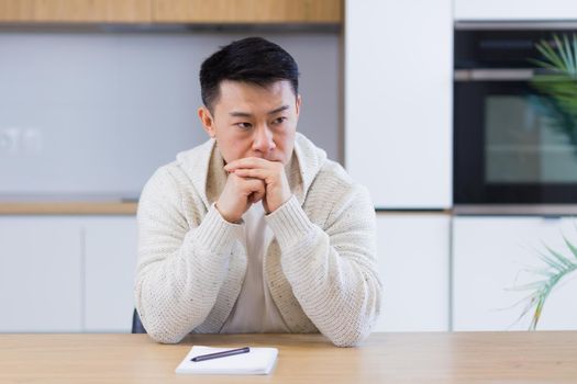 Thoughtful serious young asian man looking away. student writer sit at home office desk with laptop thinking of inspiration search solution ideas lost in thoughts dreaming solving financial problems