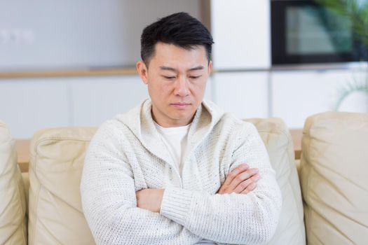 Bored friends young asian man sitting on sofa at home alone. Misses male sitting on the couch in apartment. Lonely guy indoors living room. Worried and stressed about problems in the family or at work