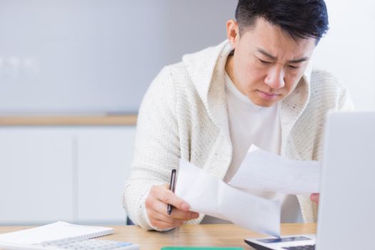 Young Asian man doing paperwork at home sitting at a desk, working on a laptop, focused on paying bills